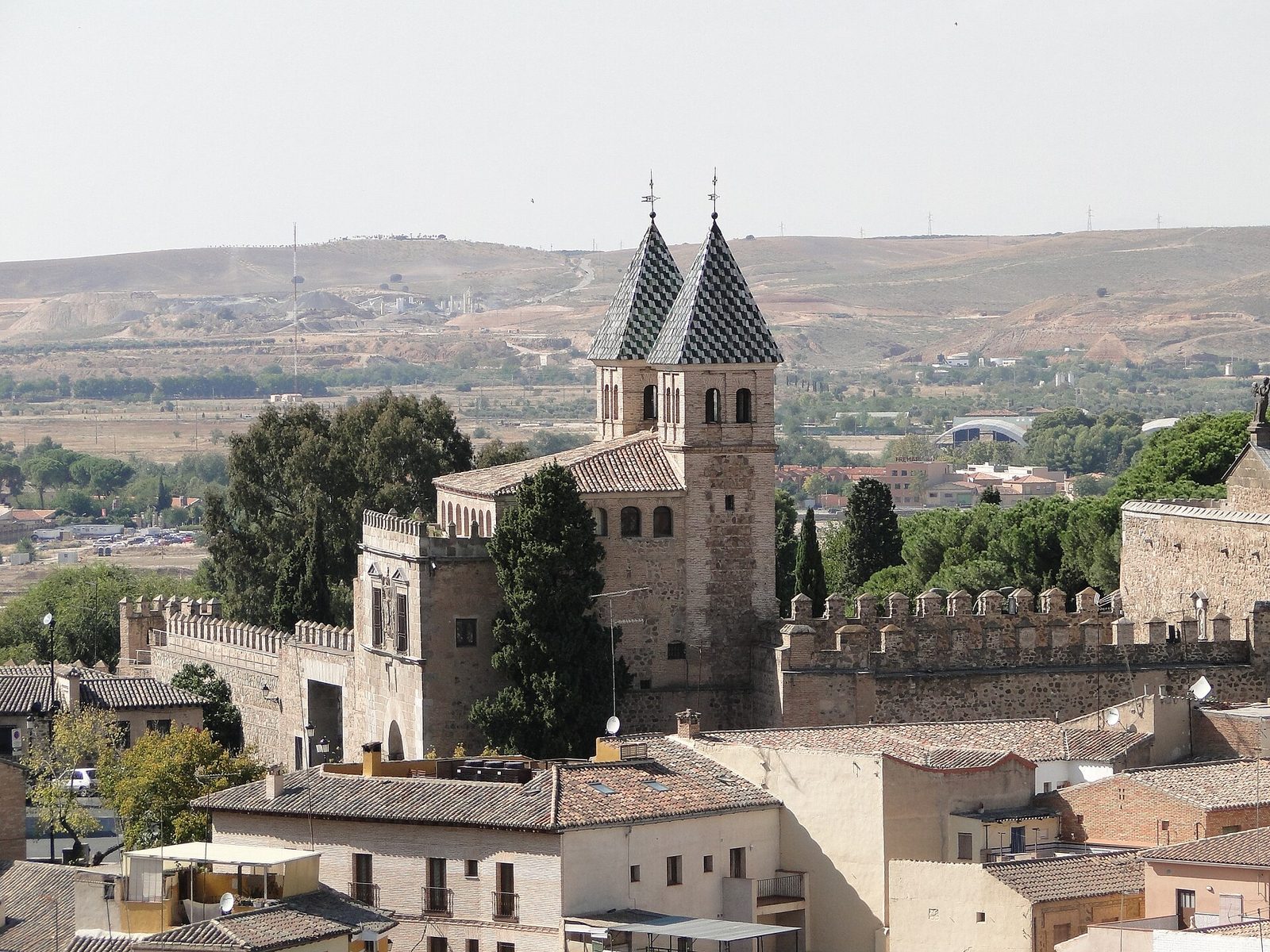 A view over Toledo's rooftops to a gate with twin towers and a stretch of walls, a church tower behind, and dry hills beyond.