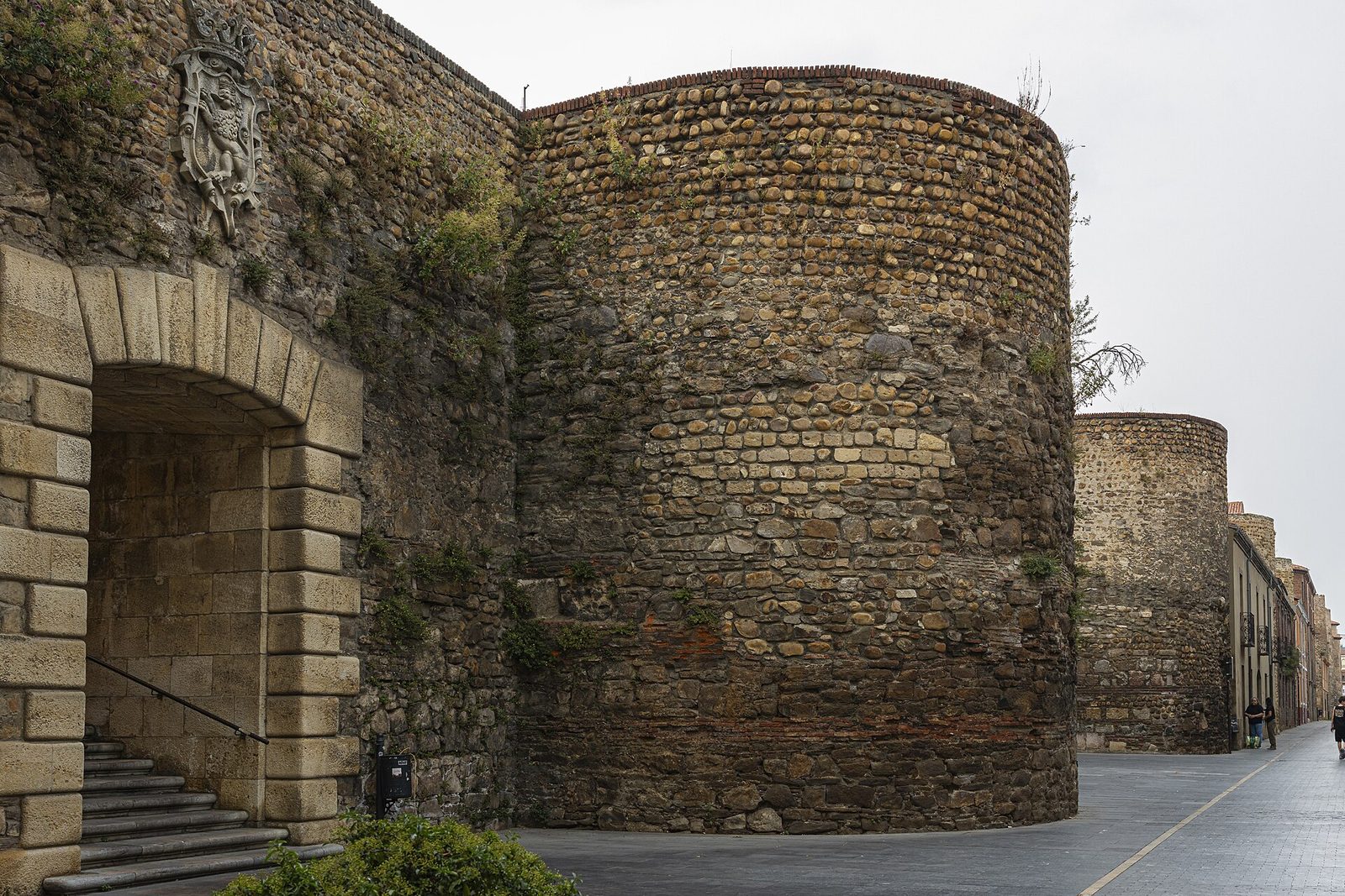 A massive wall of rough stone with a round tower, beside a paved square.