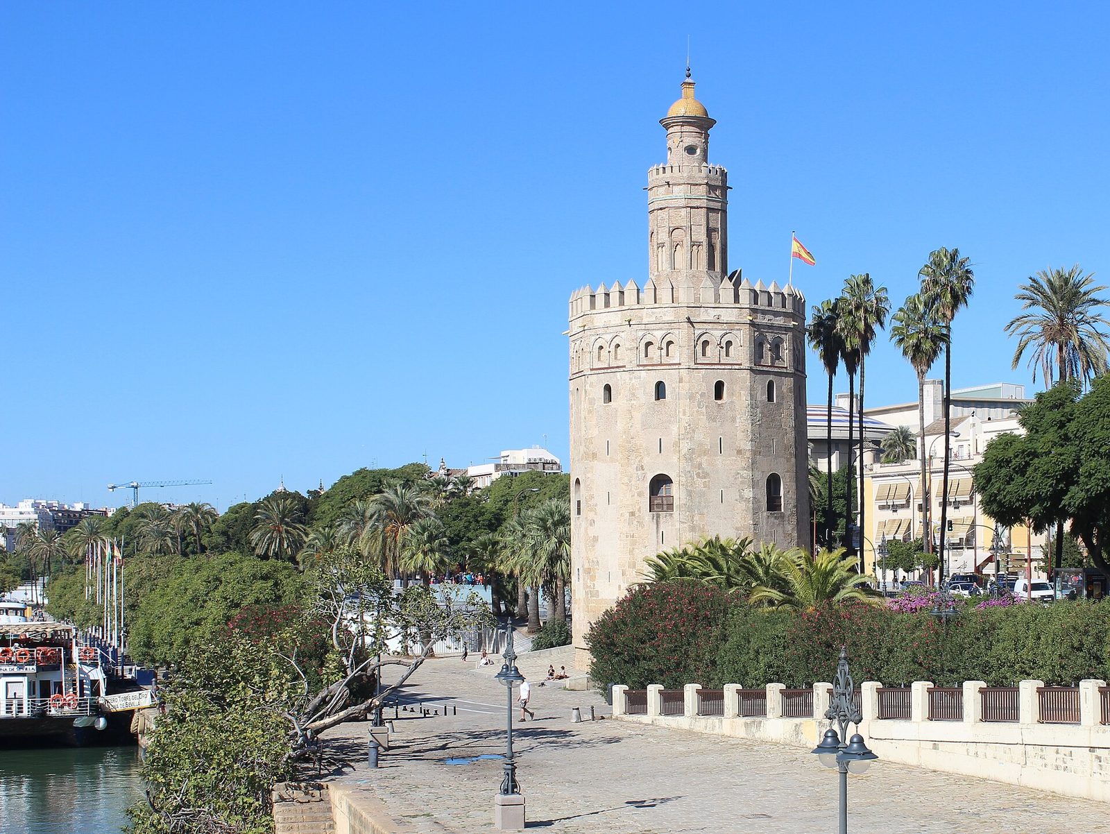 A twelve-sided stone tower with a smaller round upper stage, standing on a riverside promenade lined with palms.