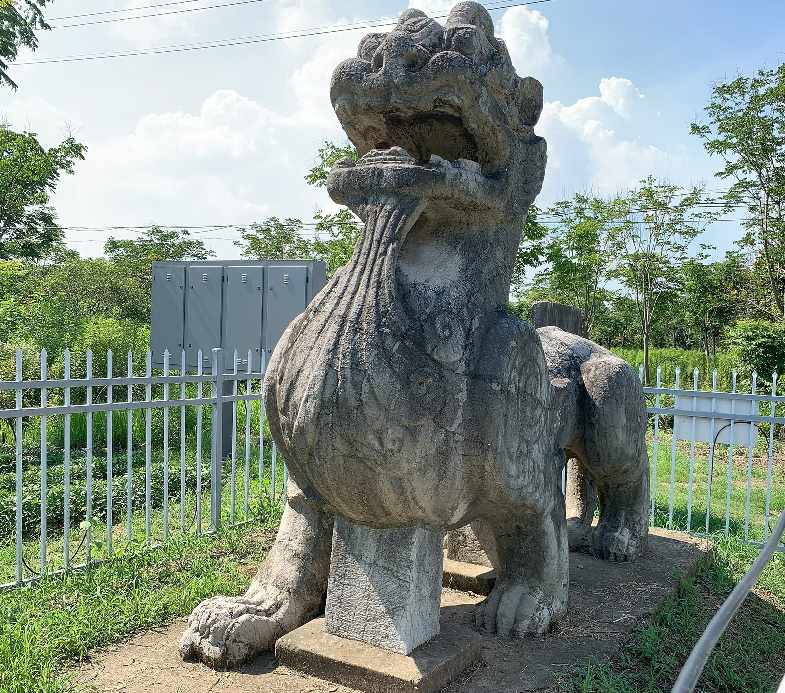 A weathered winged stone beast with a gaping mouth behind a low railing among trees.