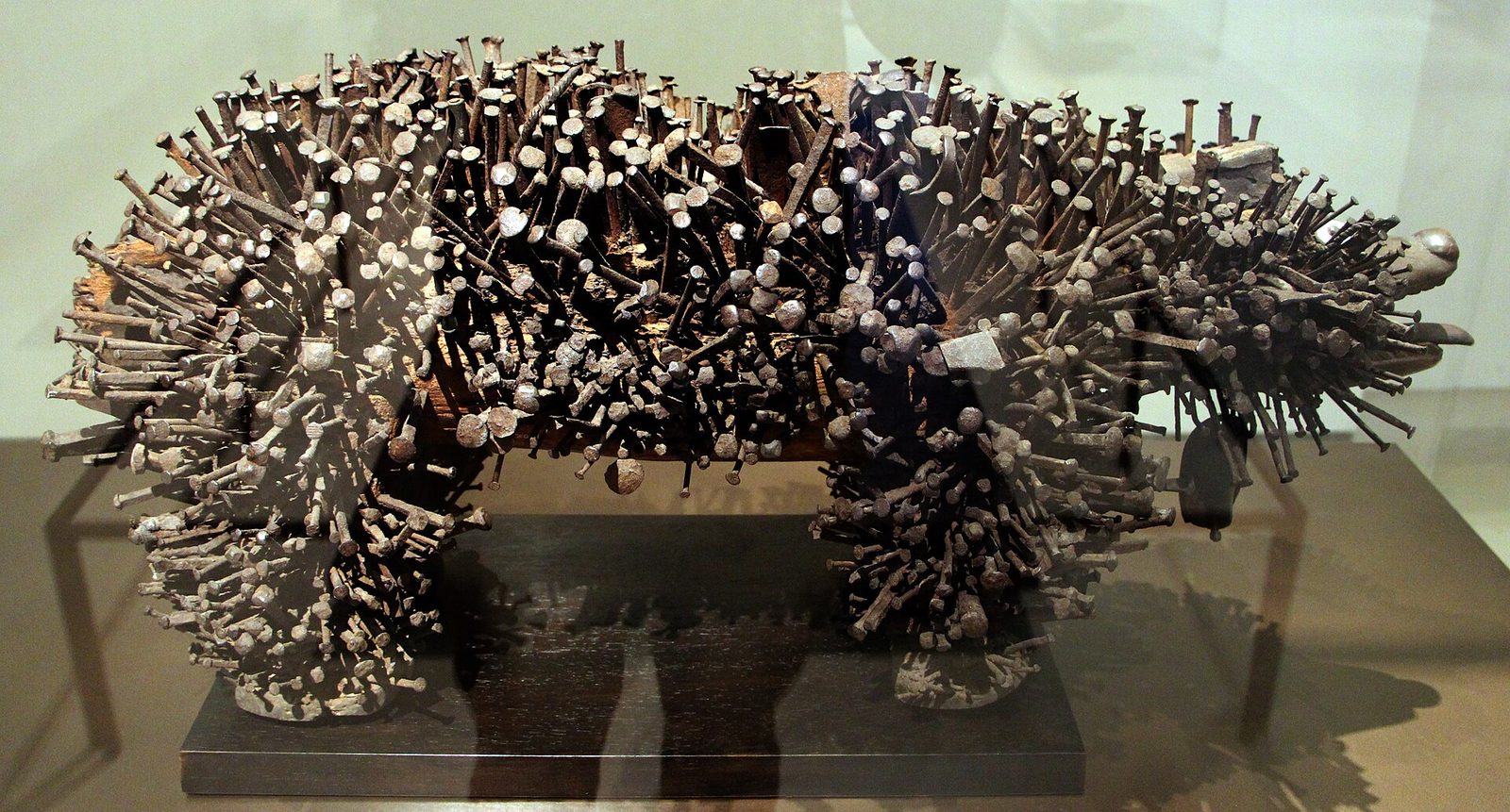 A wooden figure of a dog-like animal covered entirely in driven nails and blades, in a museum case.