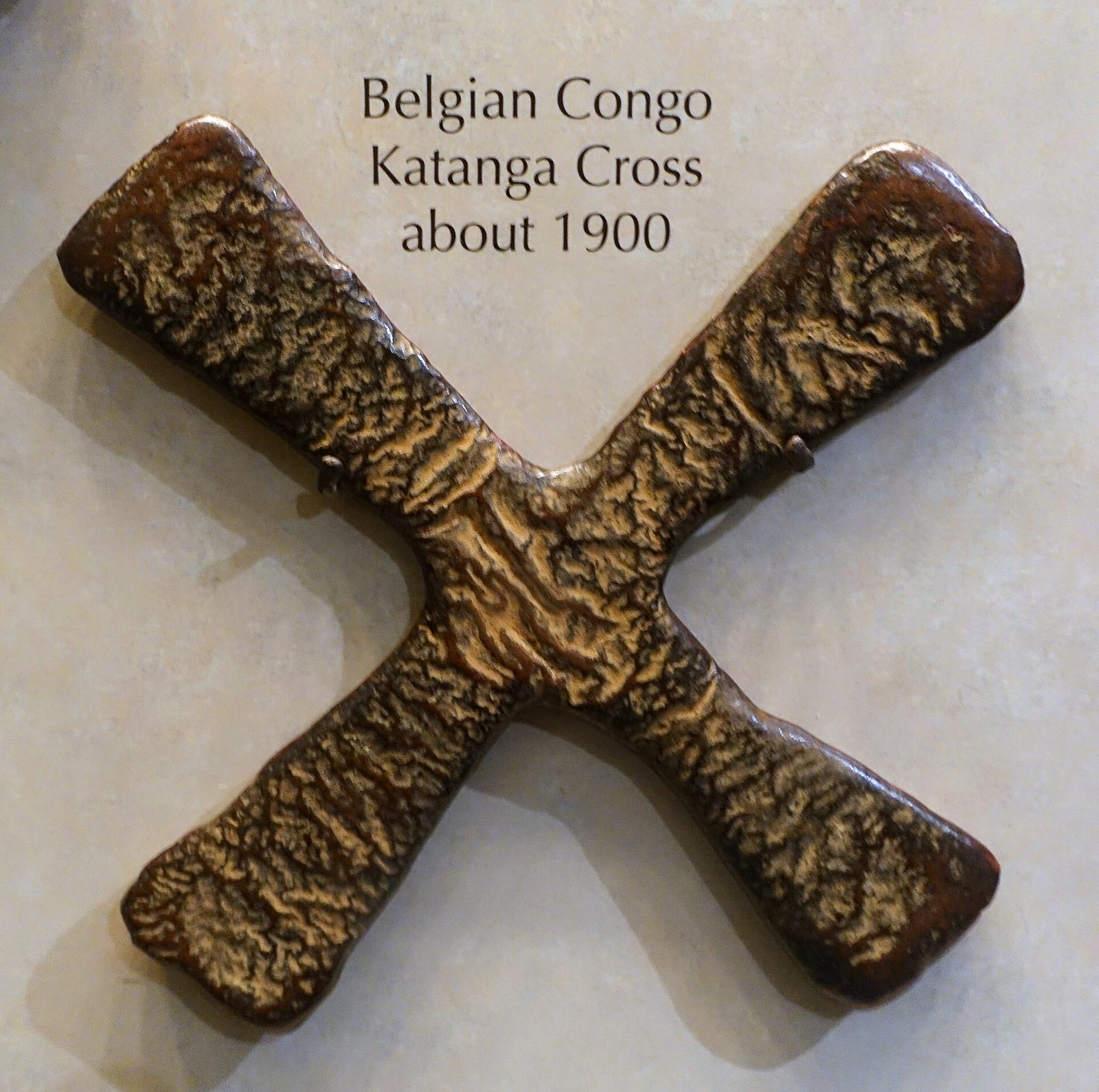 A large cast copper ingot in the shape of an X with a rough surface, on a museum wall with a label above.