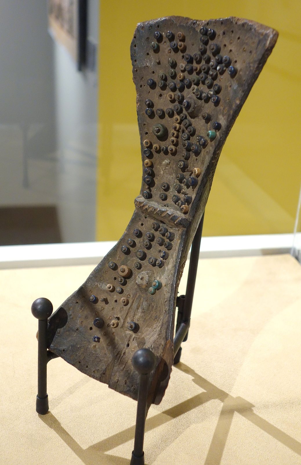 A narrow waisted wooden board studded with beads and shells, standing on a museum mount.