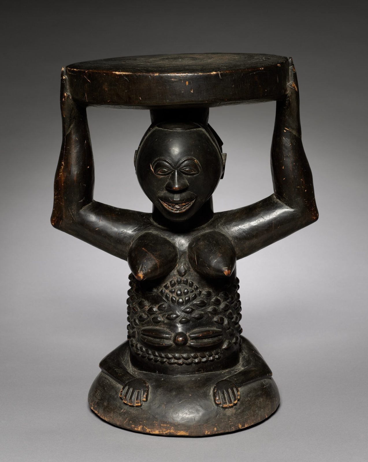 A dark wooden stool whose round seat is held up on the raised hands of a kneeling female figure with scarified skin.