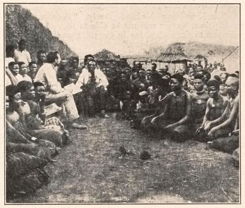 An old photograph of a European man seated among a large crowd of people sitting on the ground before huts.