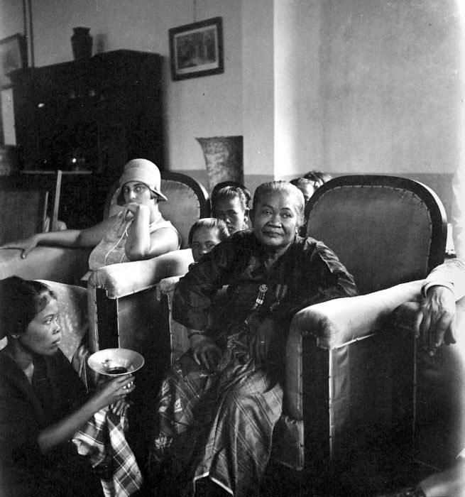 A photograph of an older woman in a dark dress seated in an armchair in a furnished room, younger family members gathered around her.
