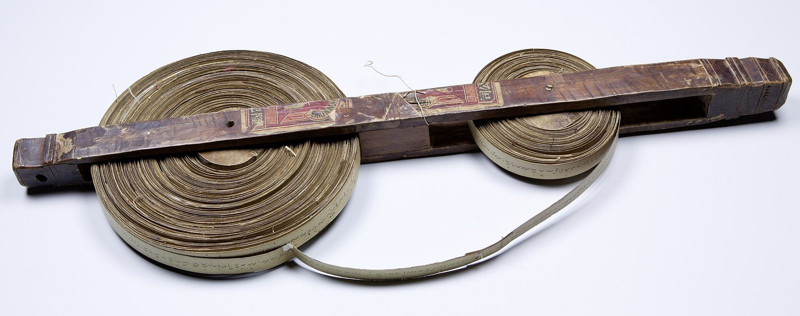 A long narrow strip of palm leaf written in fine script, wound onto two wooden spools like a tape, lying on a white ground.