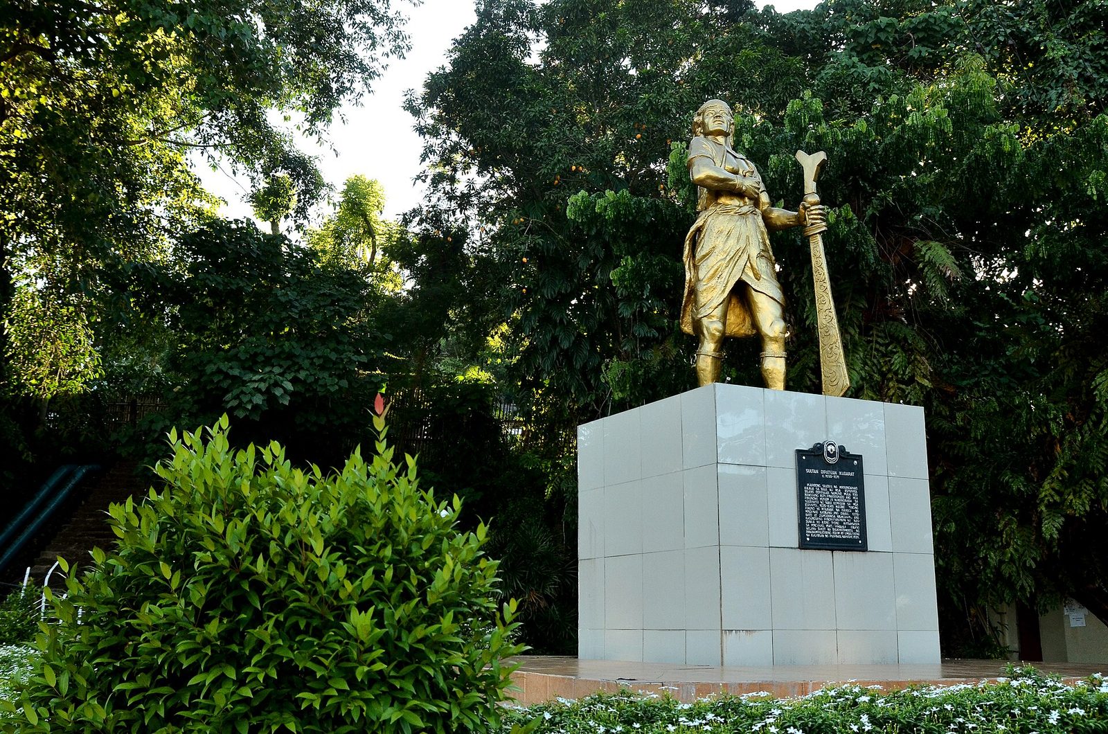 A gilded statue of a standing warrior holding a sword, on a white pedestal among trees.