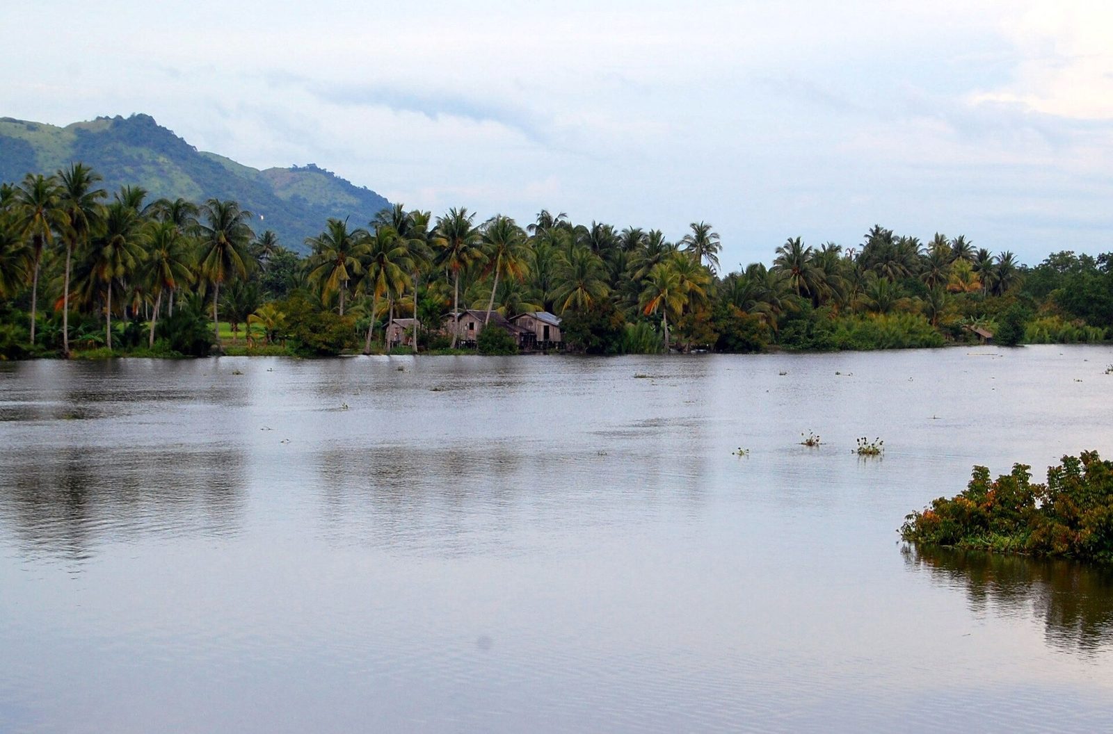 A broad calm river between low wooded banks, with stilt houses along the far shore and mountains behind.