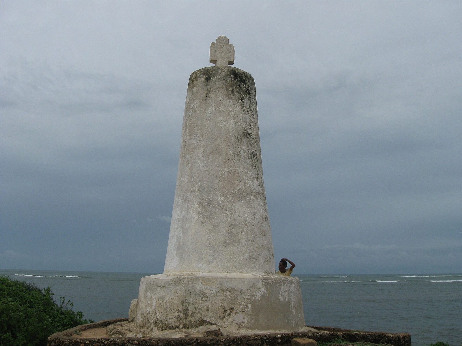 A tall whitewashed stone pillar topped with a small cross, on a headland above the sea.