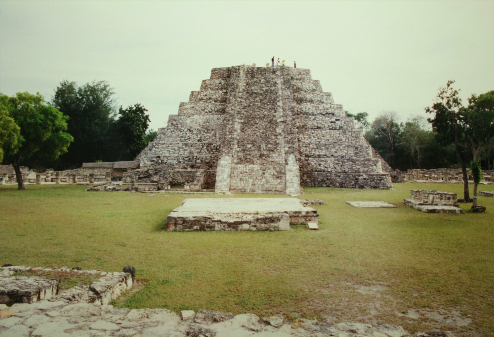 A stepped stone pyramid with a central stair in a grassy plaza, low ruined platforms around it.