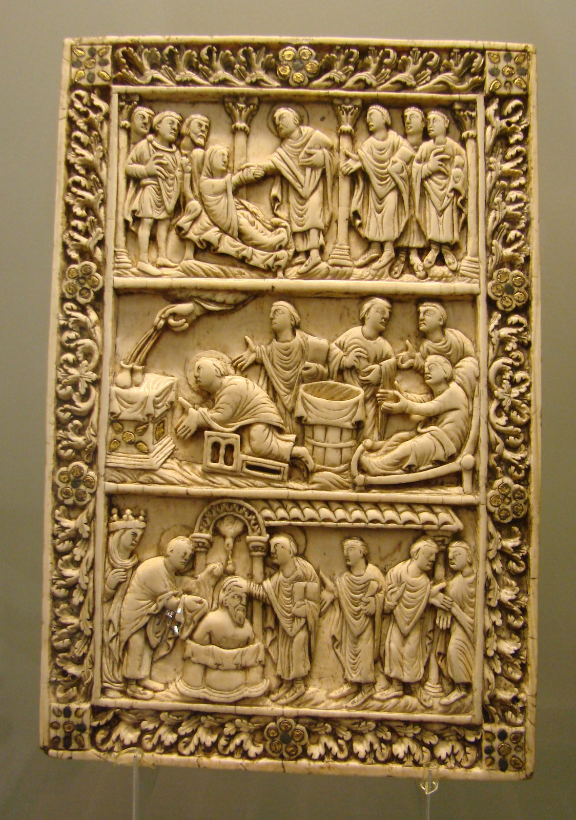 A carved ivory panel in several registers showing a naked figure standing in a round font while a bishop lays a hand on his head, with attendants and a descending dove above.