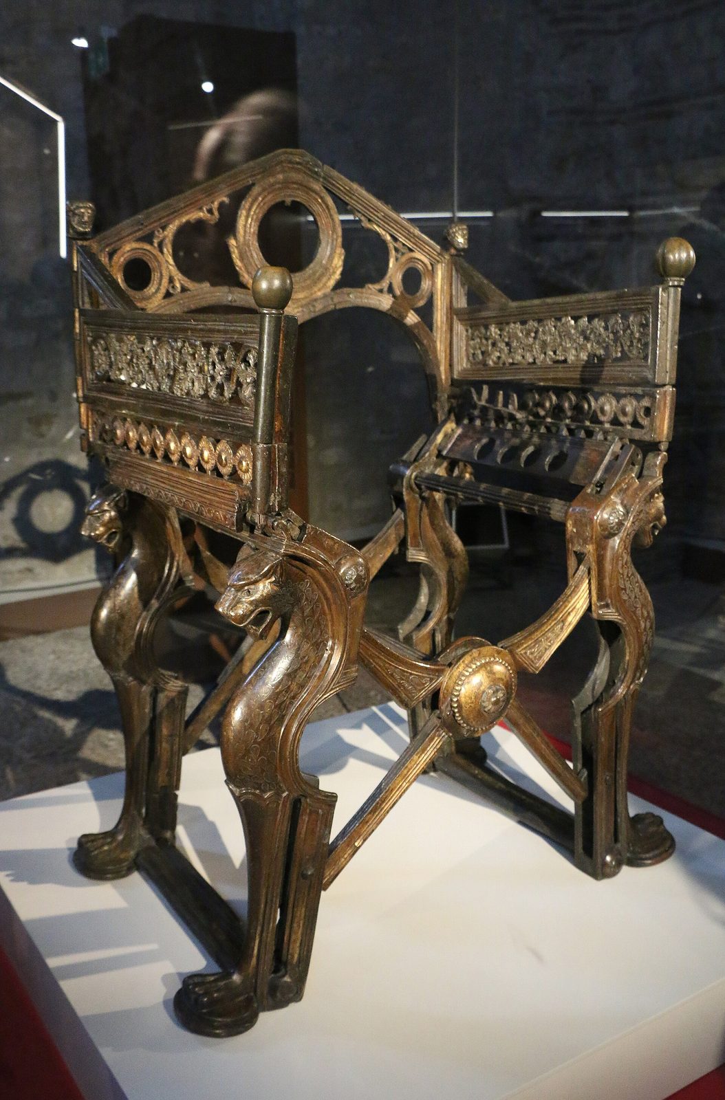A folding seat of cast bronze, its legs shaped as animal paws and heads, with a curved back and arms added above, displayed on a plinth.