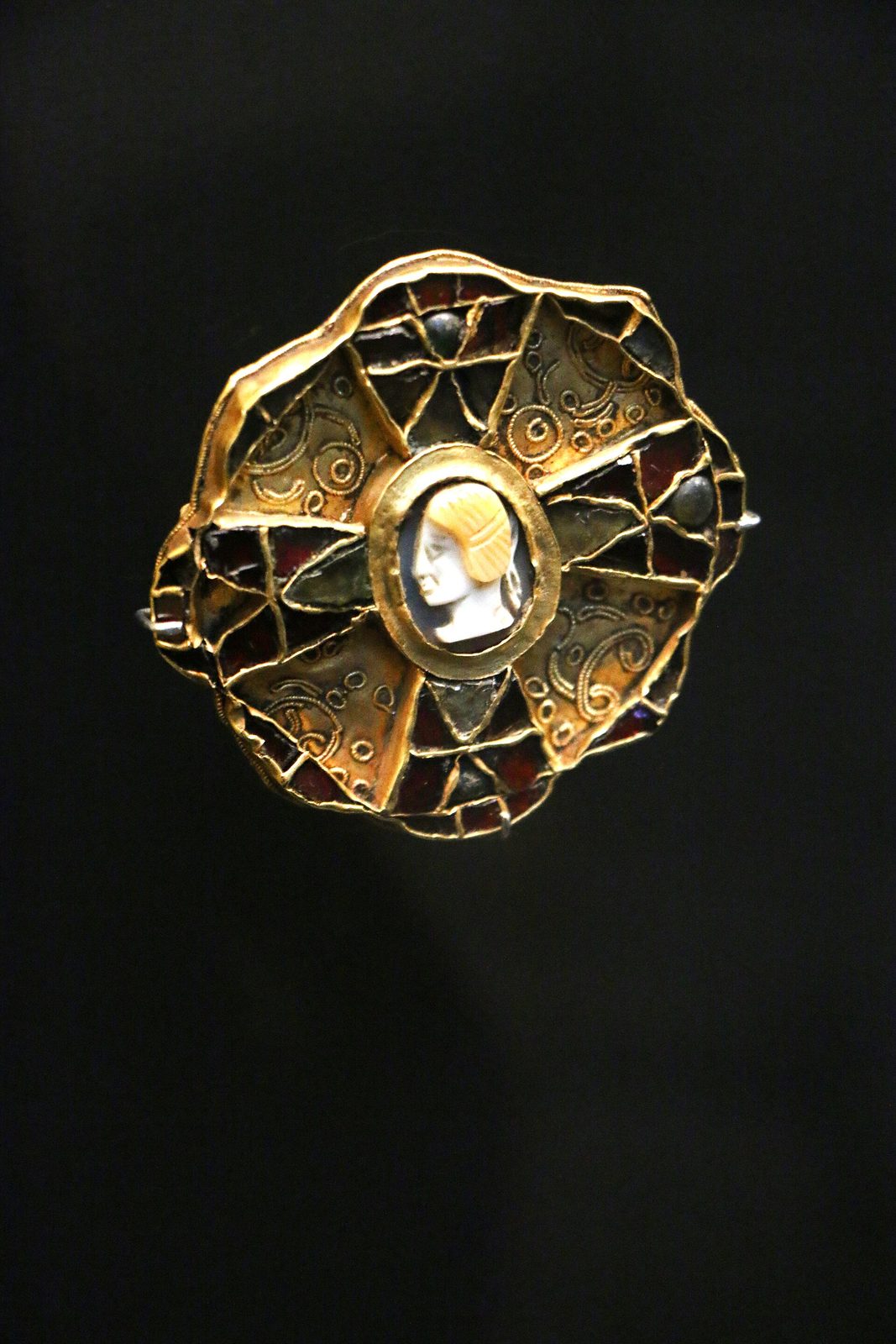 A gold brooch with a scalloped outline, its surface divided by gold cells filled with dark red garnet, and a pale carved cameo head set at the centre.
