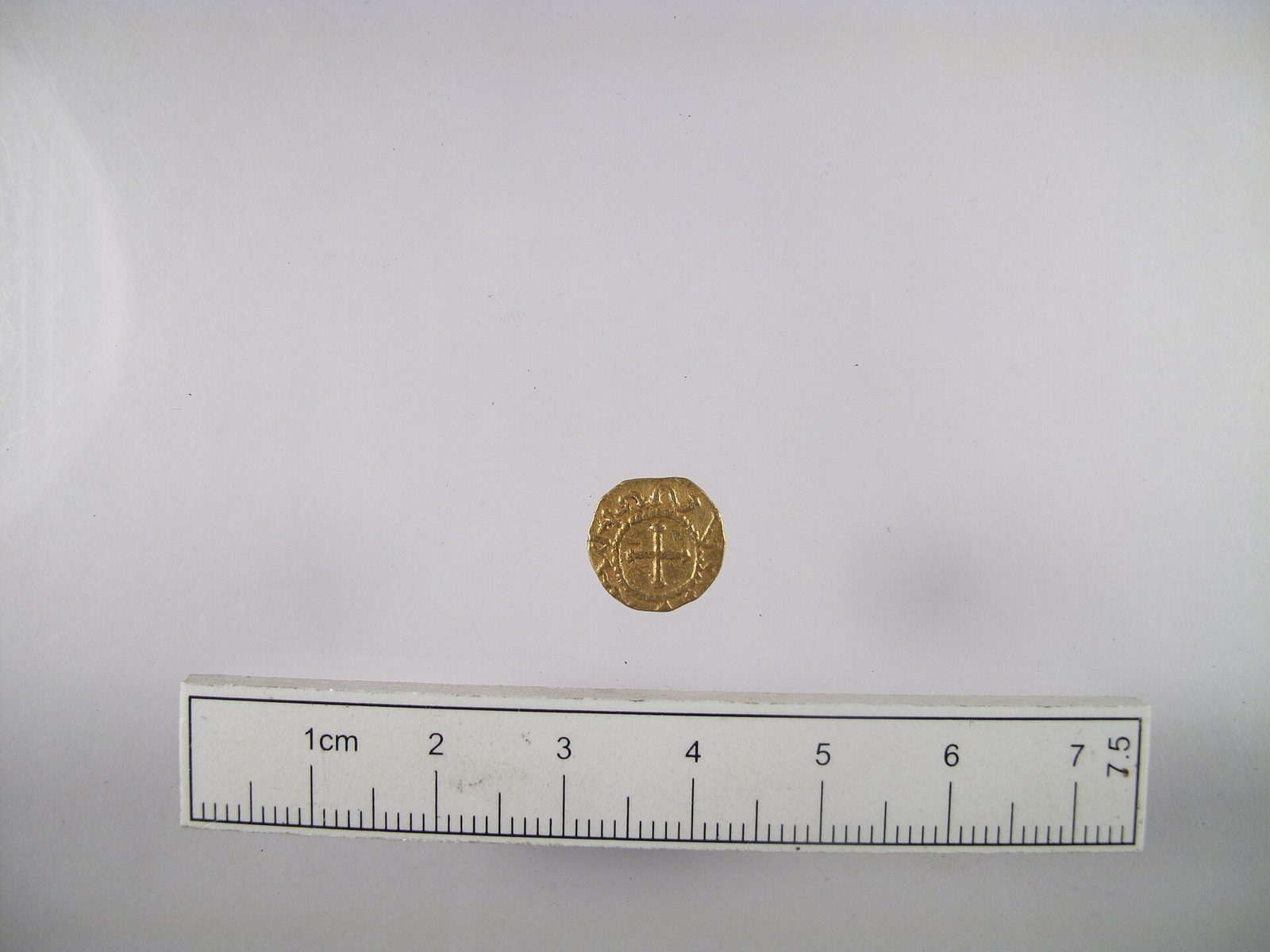 A small gold coin photographed on a plain ground beside a scale bar, its faces worn, with a facing head on one and a cross on the other.