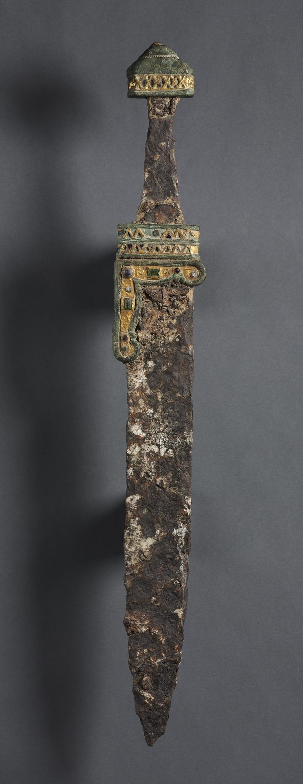 A long single-edged iron blade, corroded brown and tapering to a point, photographed upright against a plain pale ground.