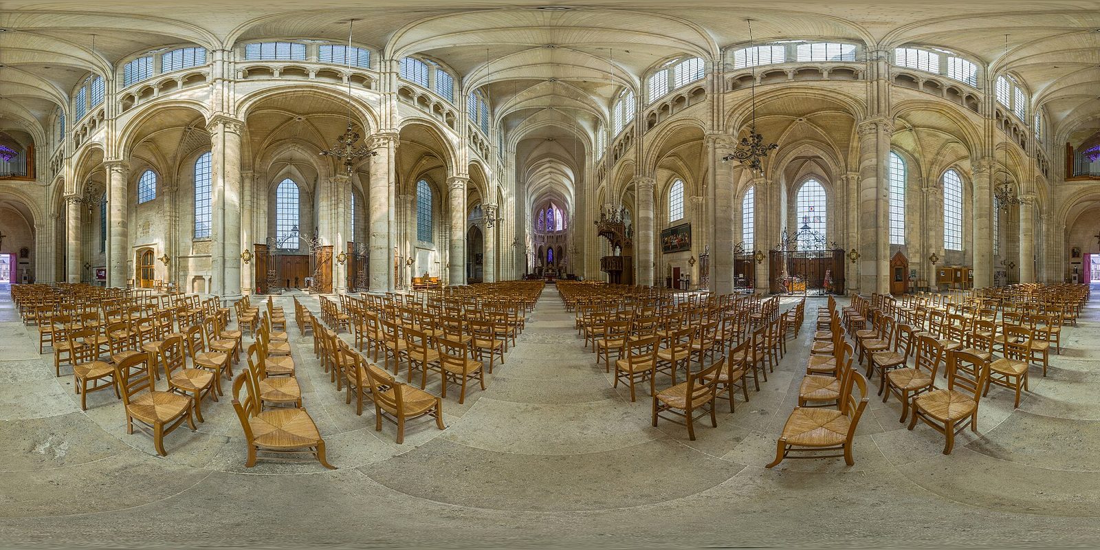 A wide panoramic view down the interior of a Gothic cathedral, tall clustered piers on both sides carrying a high vault, with rows of chairs on the stone floor.