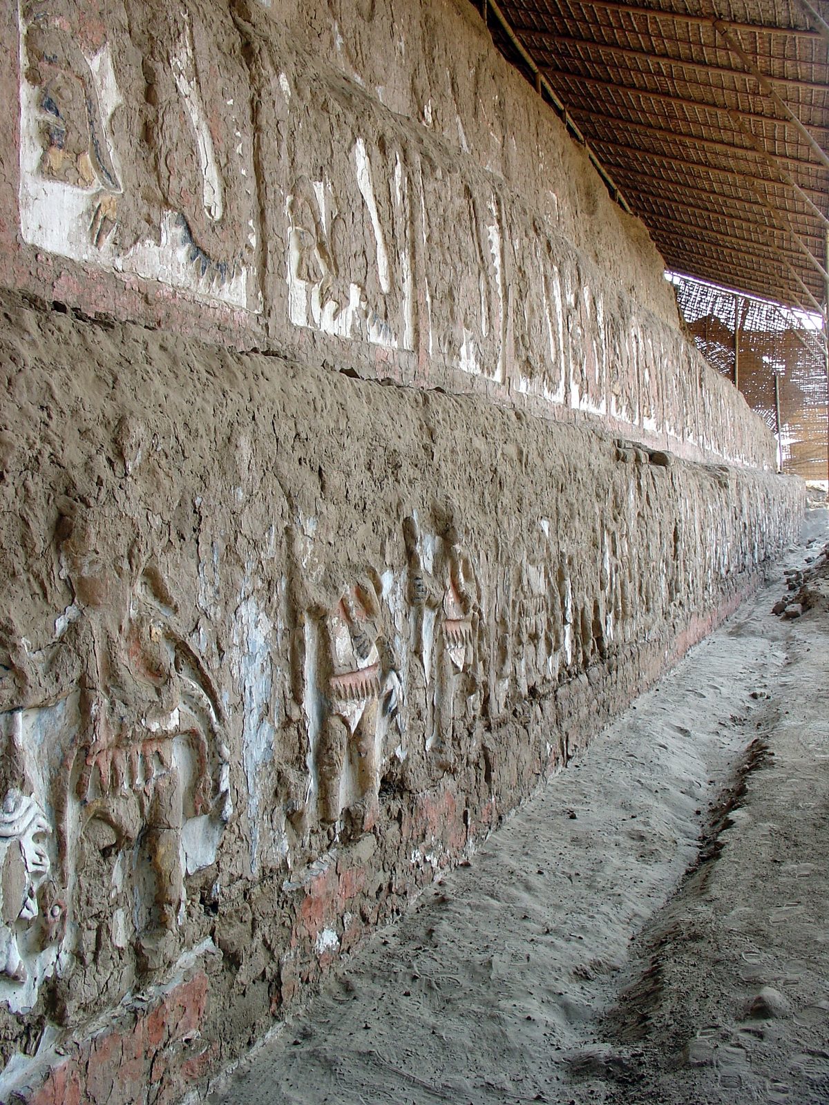A long mud wall carved with rows of painted relief figures in red, white and yellow, under a protective roof.
