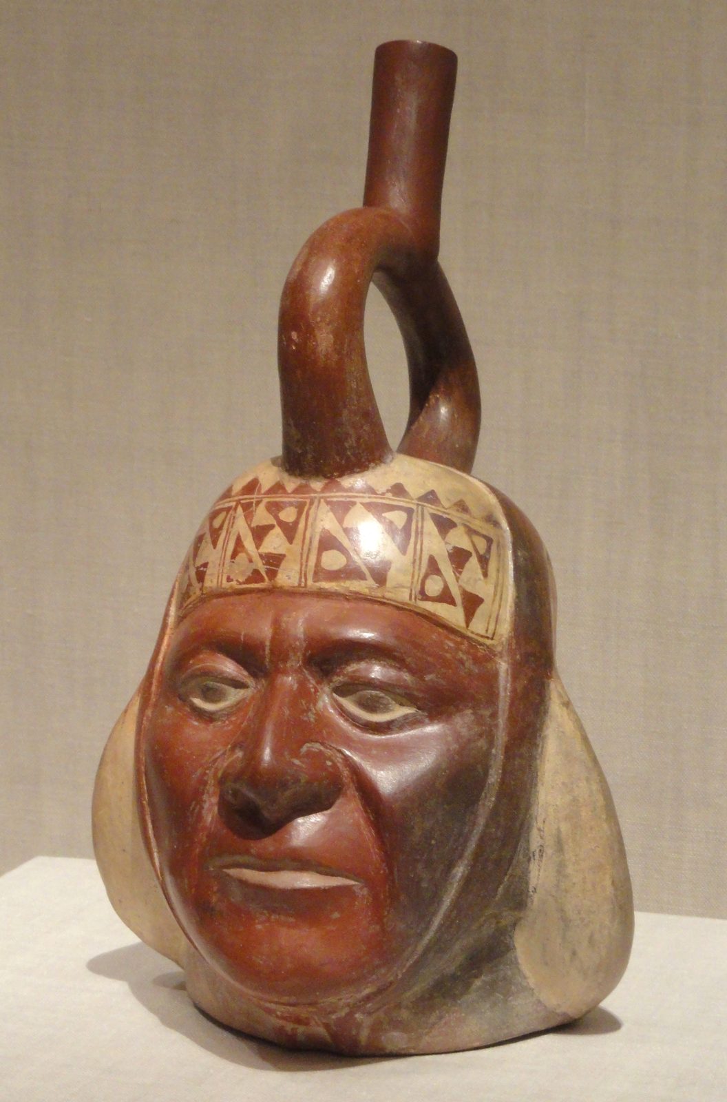 A red and cream ceramic stirrup-spout vessel modelled as a man's head in a patterned headdress.