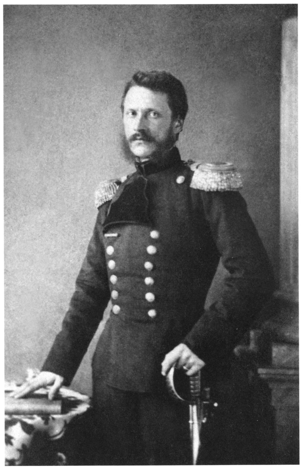 A black-and-white photograph of a moustached man in a buttoned military tunic, one hand on a sword.