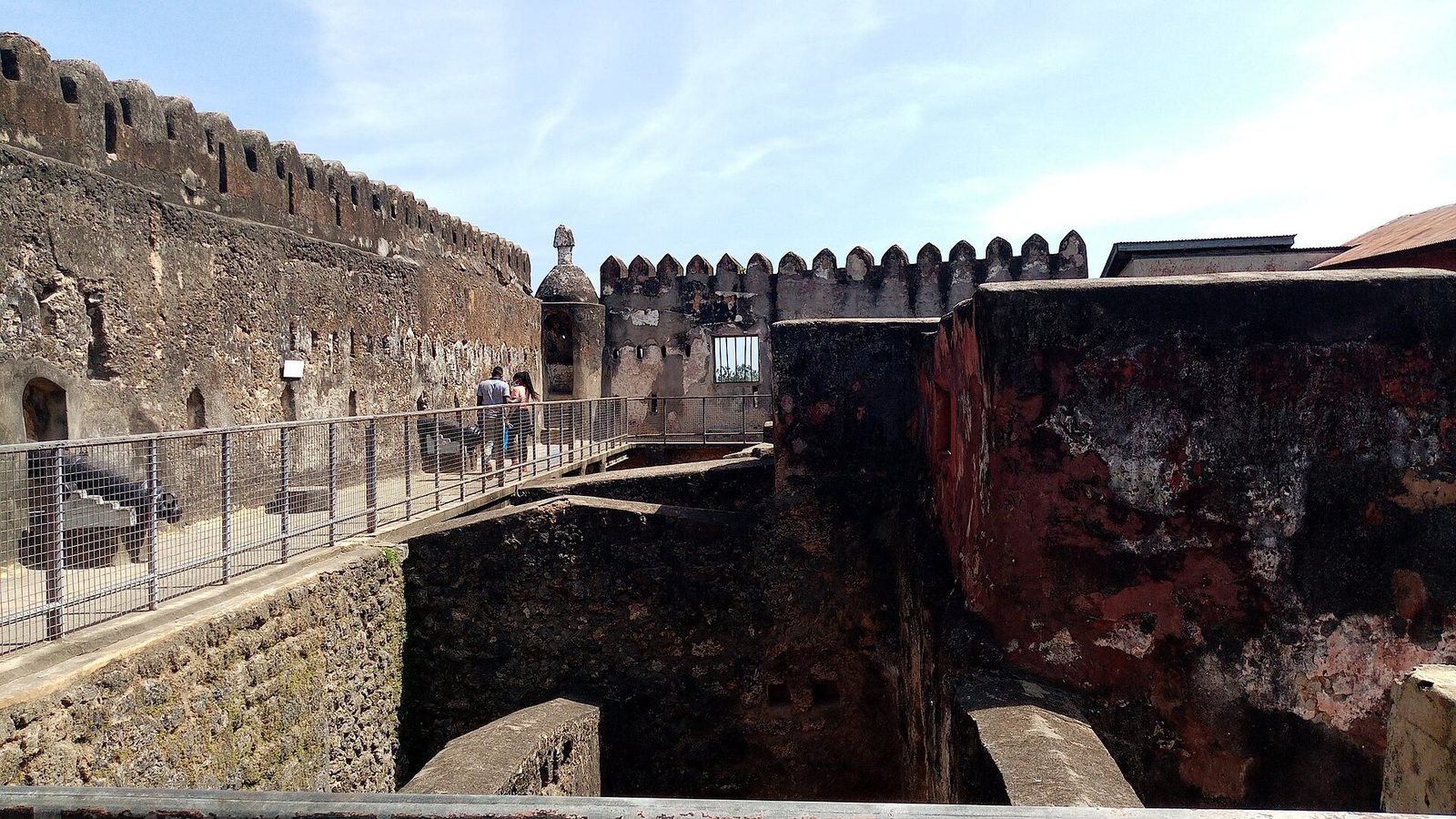 Weathered stone ramparts with crenellations and a walkway inside an old fort, under a pale sky.