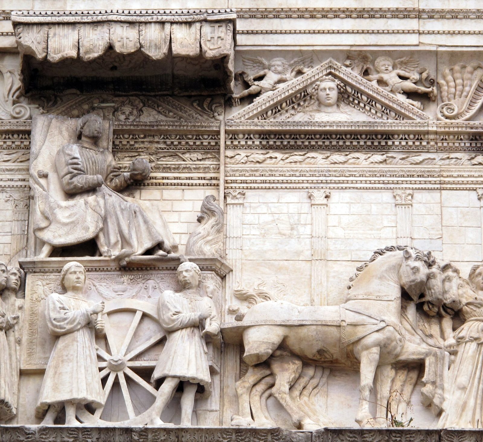 A carved marble relief: a king in armour sits enthroned under a fringed canopy on a triumphal car, attendants walking at the wheel below him, and a team of horses drawing the car away to the right.