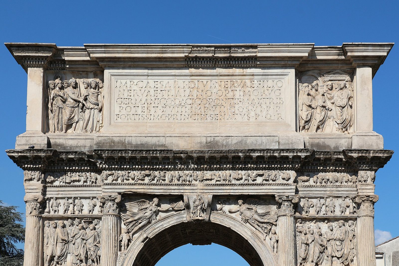 A single-arched Roman gateway of pale marble standing free in a paved street, its faces covered with relief panels of figures and processions and a long inscription across the attic.