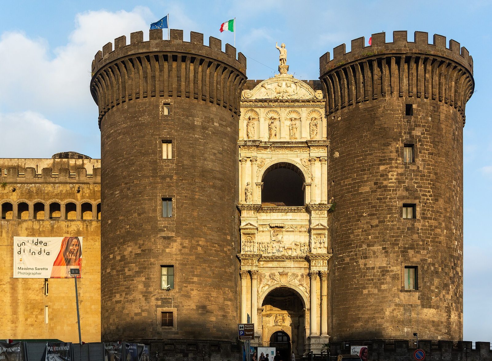 A castle of dark stone with five massive round towers and a crenellated parapet, and set between two of the towers a tall white marble gateway of stacked classical arches and reliefs.