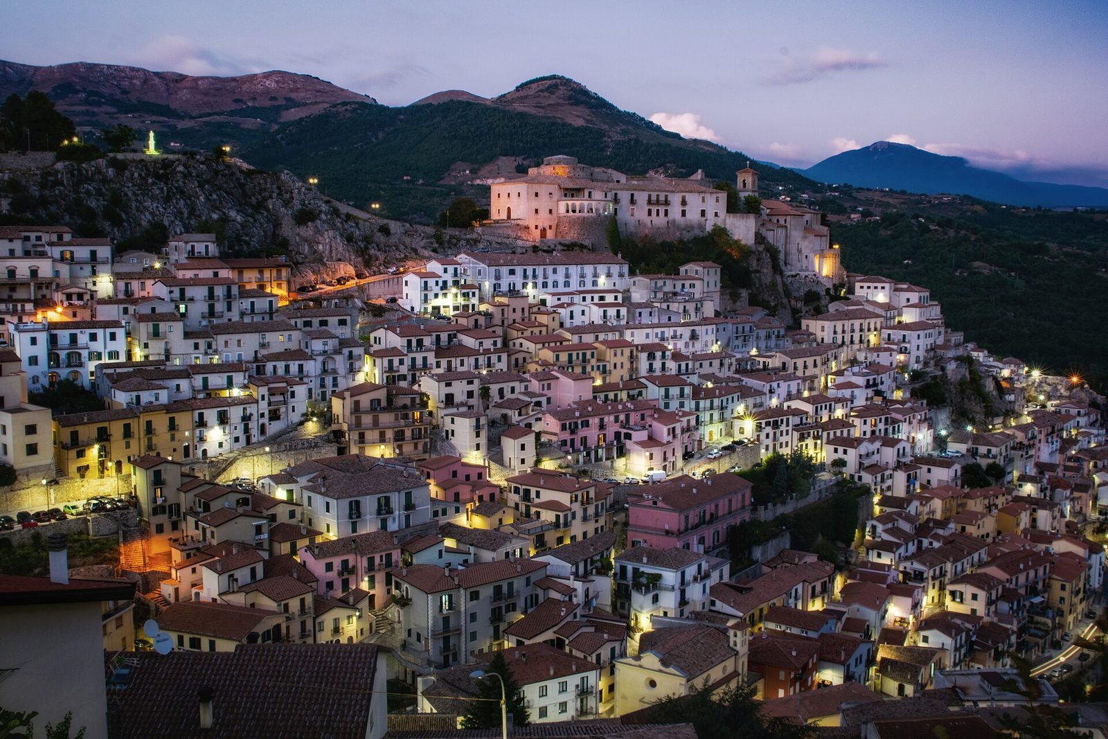 A hill town at dusk, its pale houses stacked in close tiers up a steep ridge and lit from inside, with a church tower on the crest and dark mountains behind.