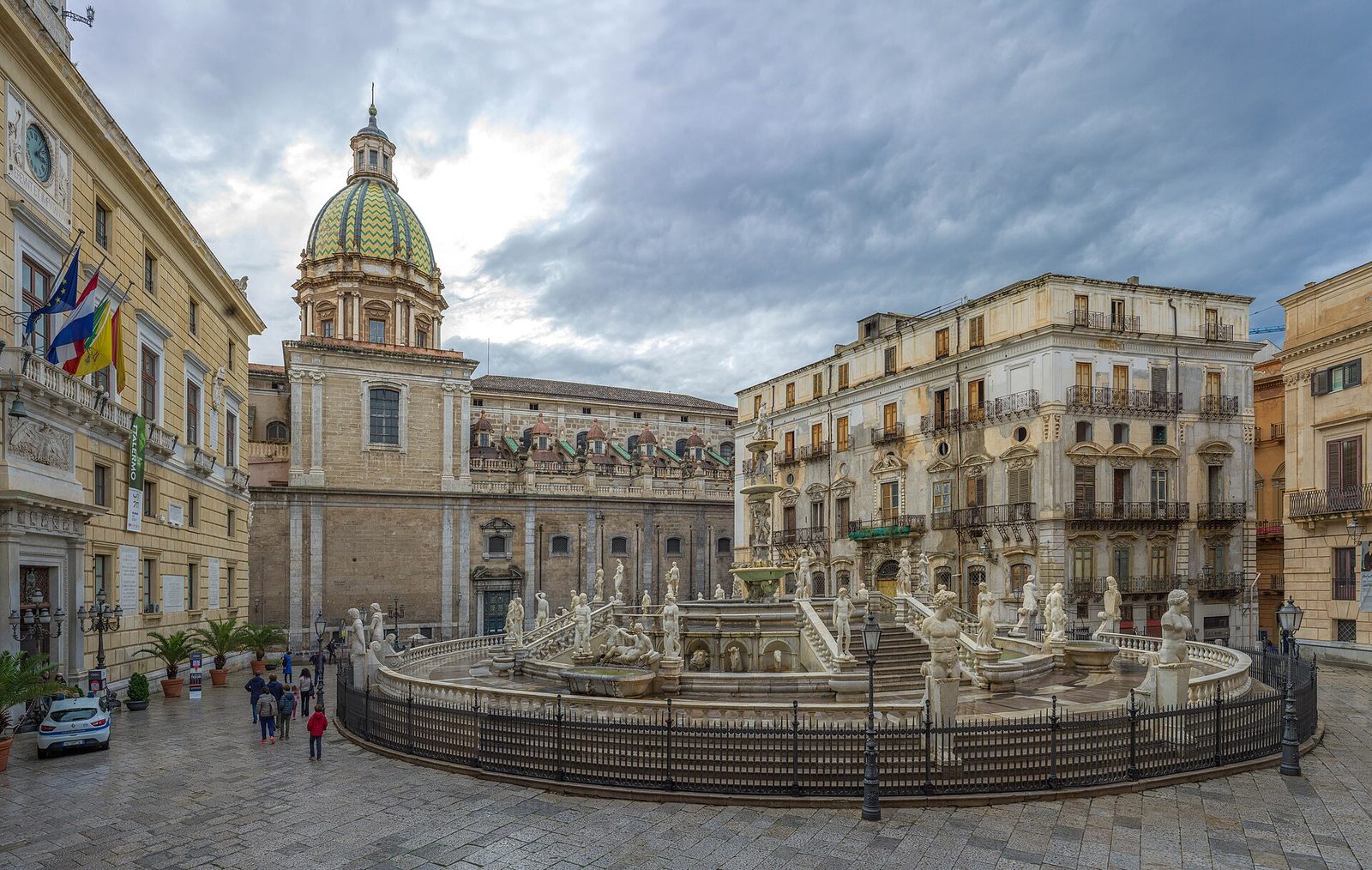 A wide paved square with a circular balustraded fountain at its centre, crowded with marble figures and basins on several levels, ringed by tall stuccoed palaces and a domed church.
