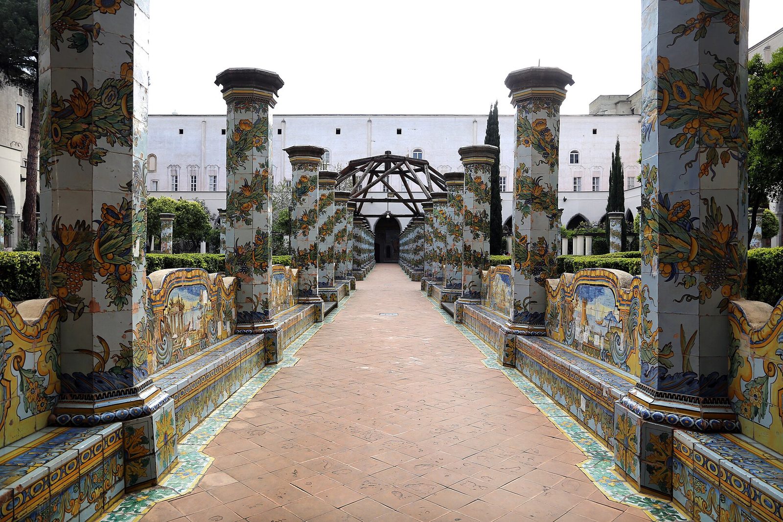 A long cloister walk paved in terracotta, running between rows of broad octagonal piers faced from top to bottom in painted tiles of yellow, blue and green, with tiled benches and a garden beyond.