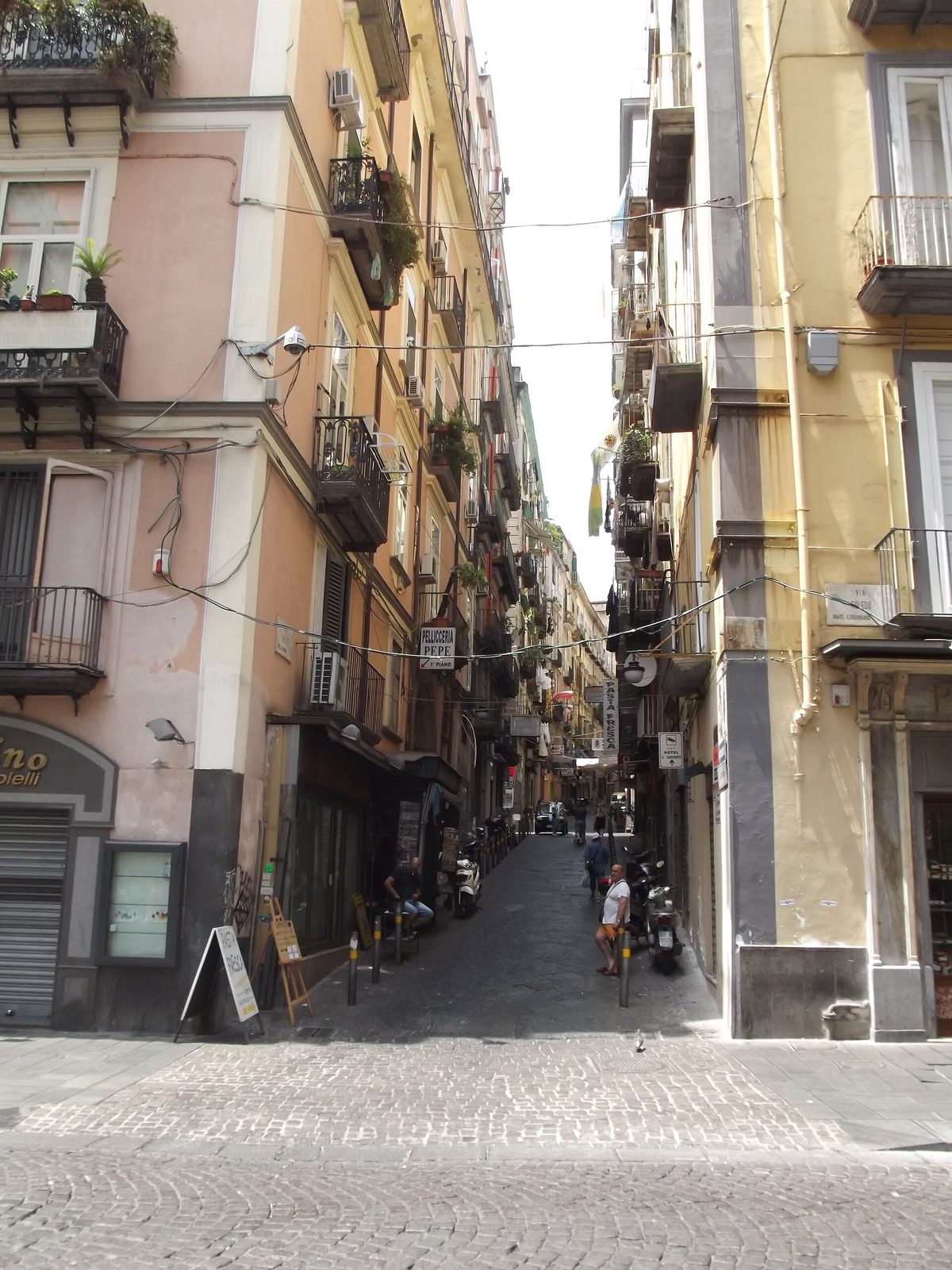 A narrow lane climbing between tall tenement blocks, washing and air conditioners on the balconies, mopeds parked along the walls and a few people on the paving at the bottom.
