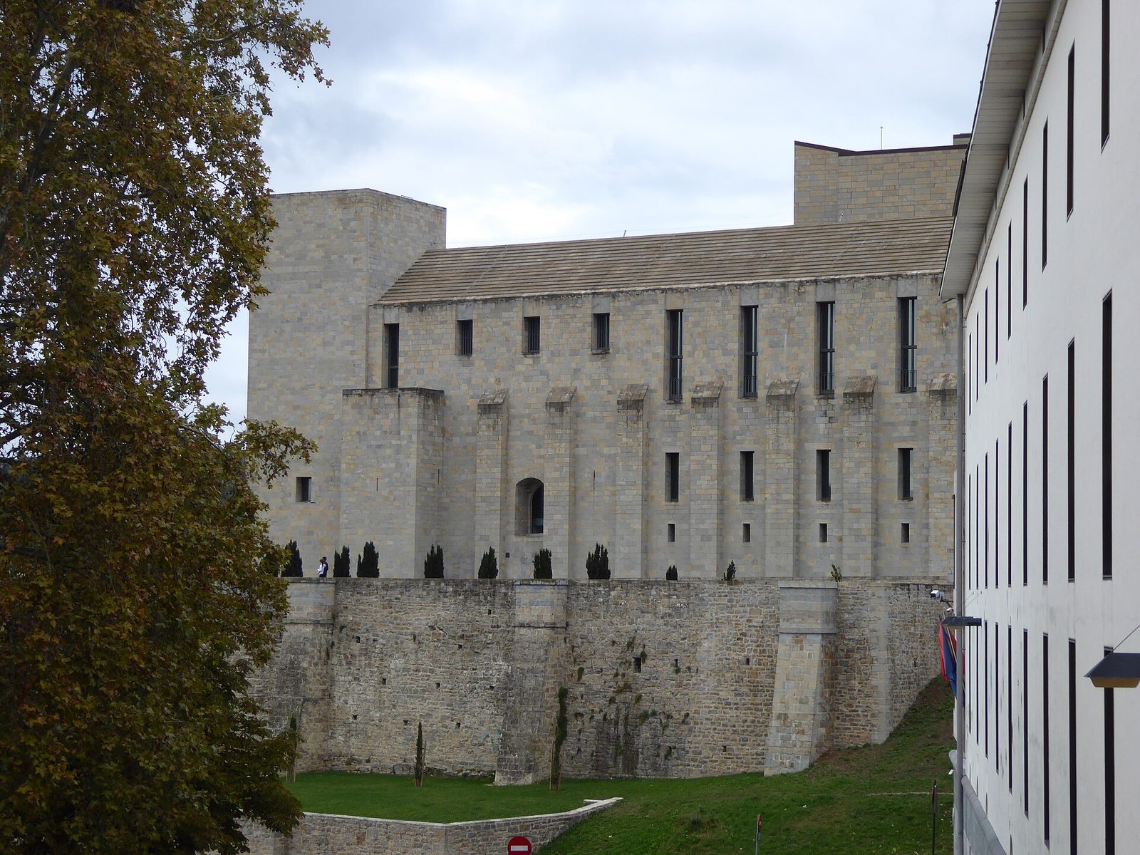 A large stone palace with plain walls and small windows beside a lawn, under a cloudy sky.