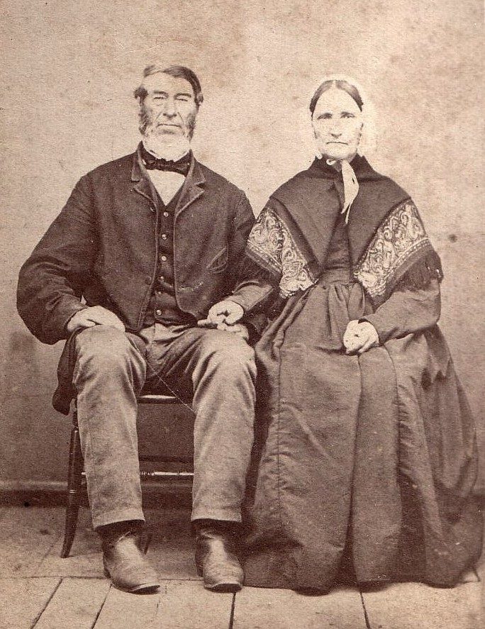 A sepia photograph of a bearded man and a woman in a bonnet and shawl, seated side by side.