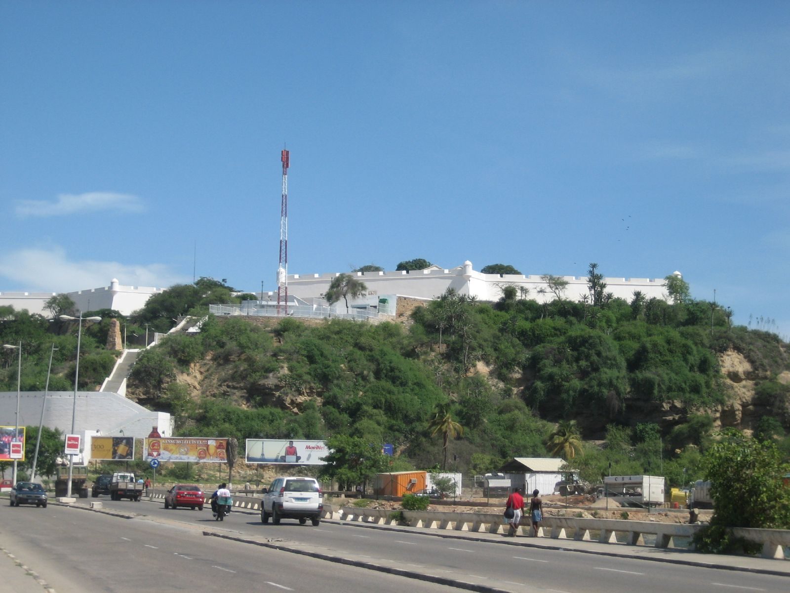 A low white fortress on a green hill above a busy coastal road.