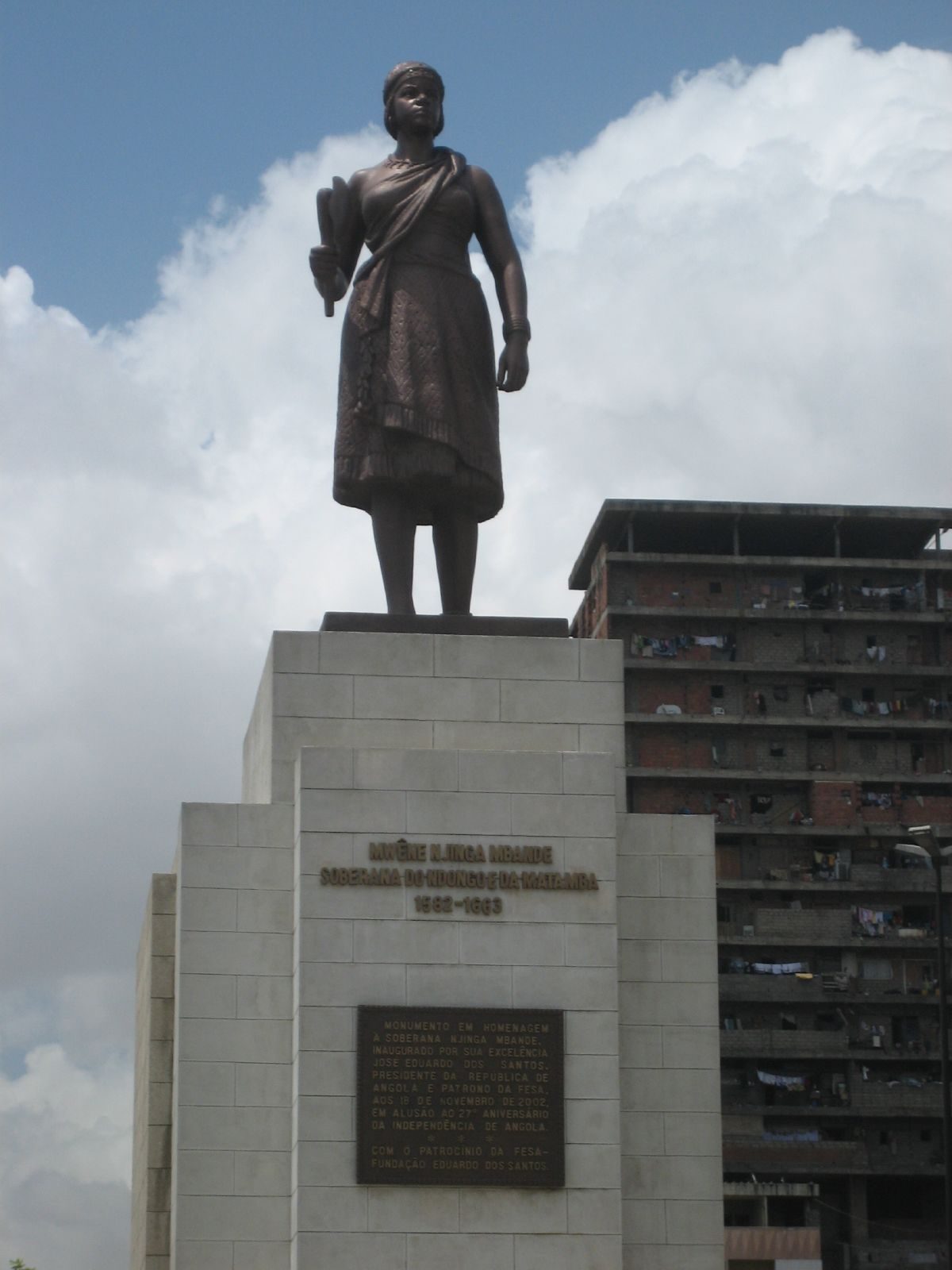 A bronze statue of a standing woman on a tall stone pedestal, tower blocks behind.