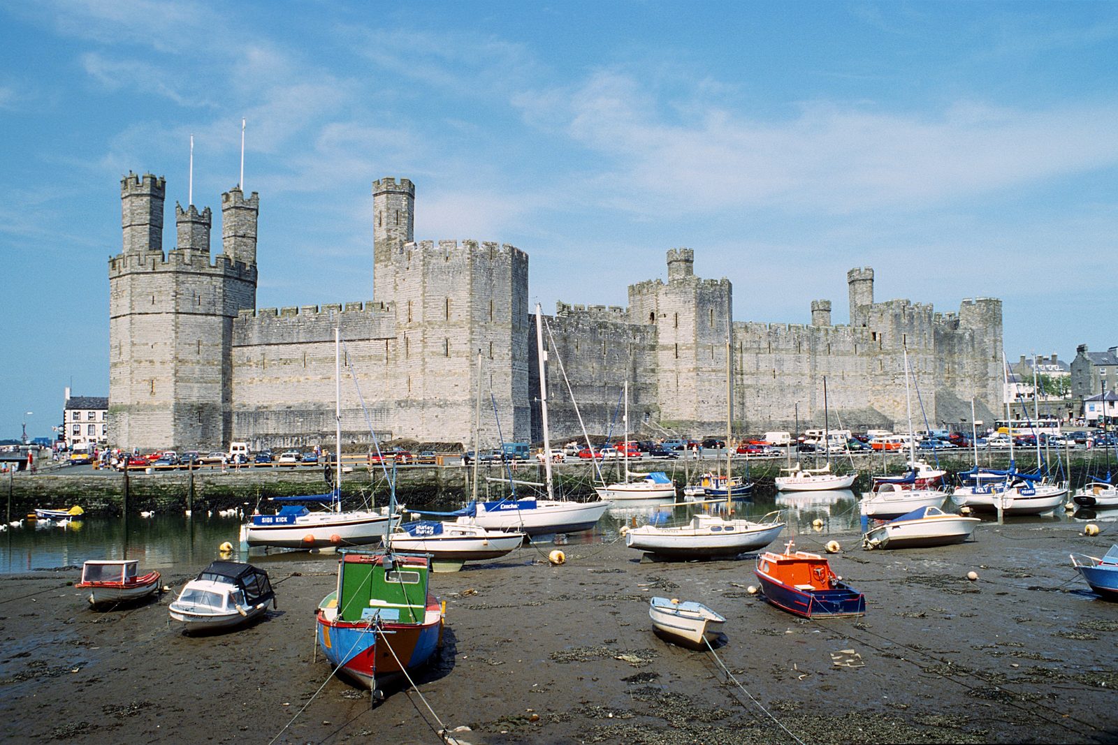 A long castle wall with polygonal towers above a harbour full of small boats at low tide.