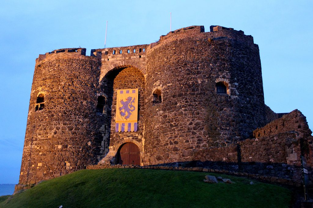 A twin-towered stone castle gatehouse lit by evening sun, a banner hanging over its arch.