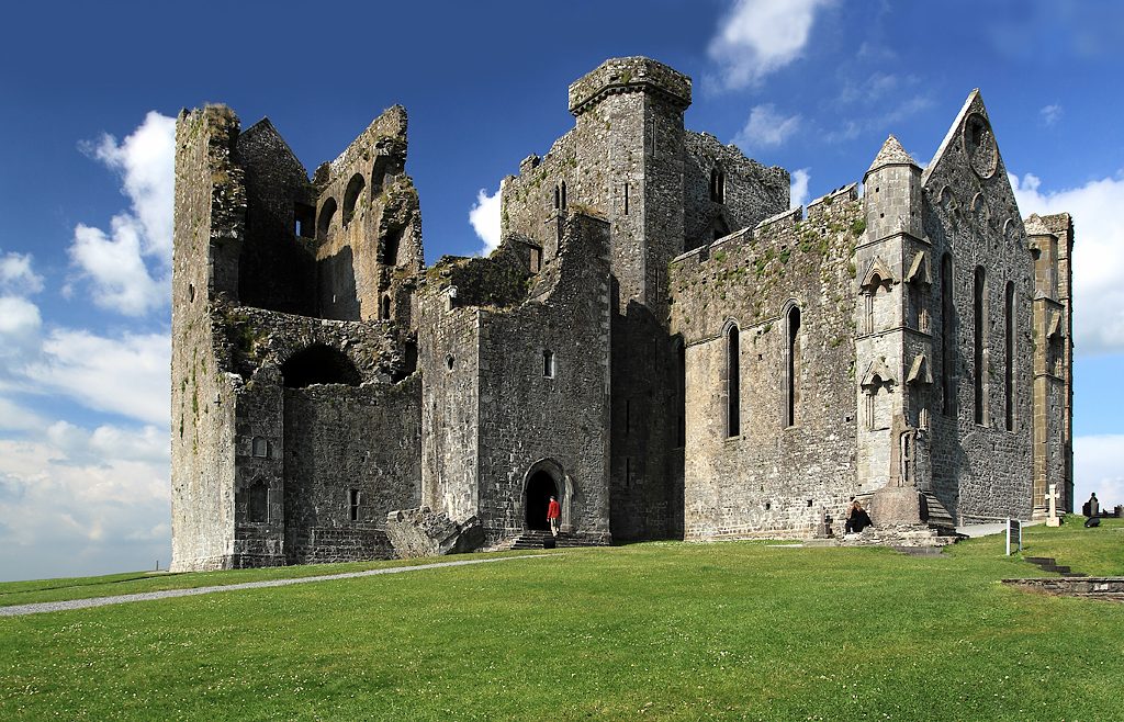 Ruined medieval church and tower buildings on a grassy rock under a blue sky.