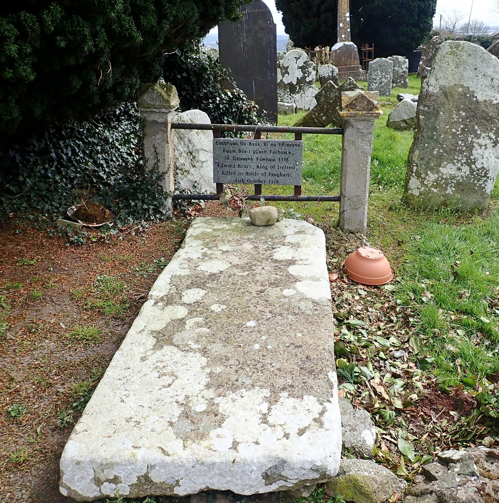 A worn stone slab lying in a churchyard, a plaque on the railing behind it.
