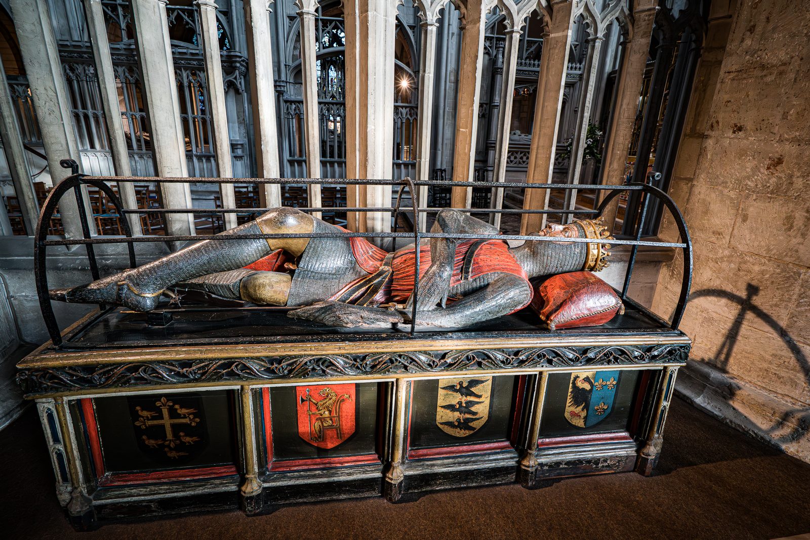 A painted wooden effigy of a knight in mail lying cross-legged on a tomb chest inside an iron frame.