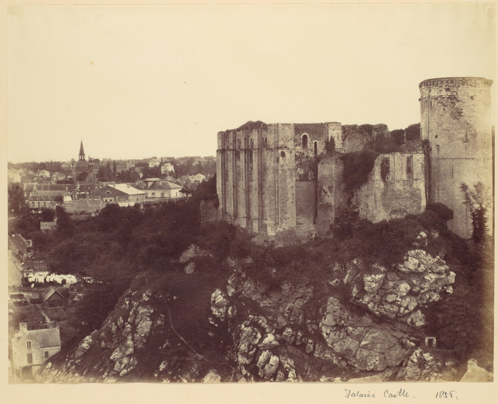 An old photograph of a ruined square keep and a round tower on a rocky crag above a town.