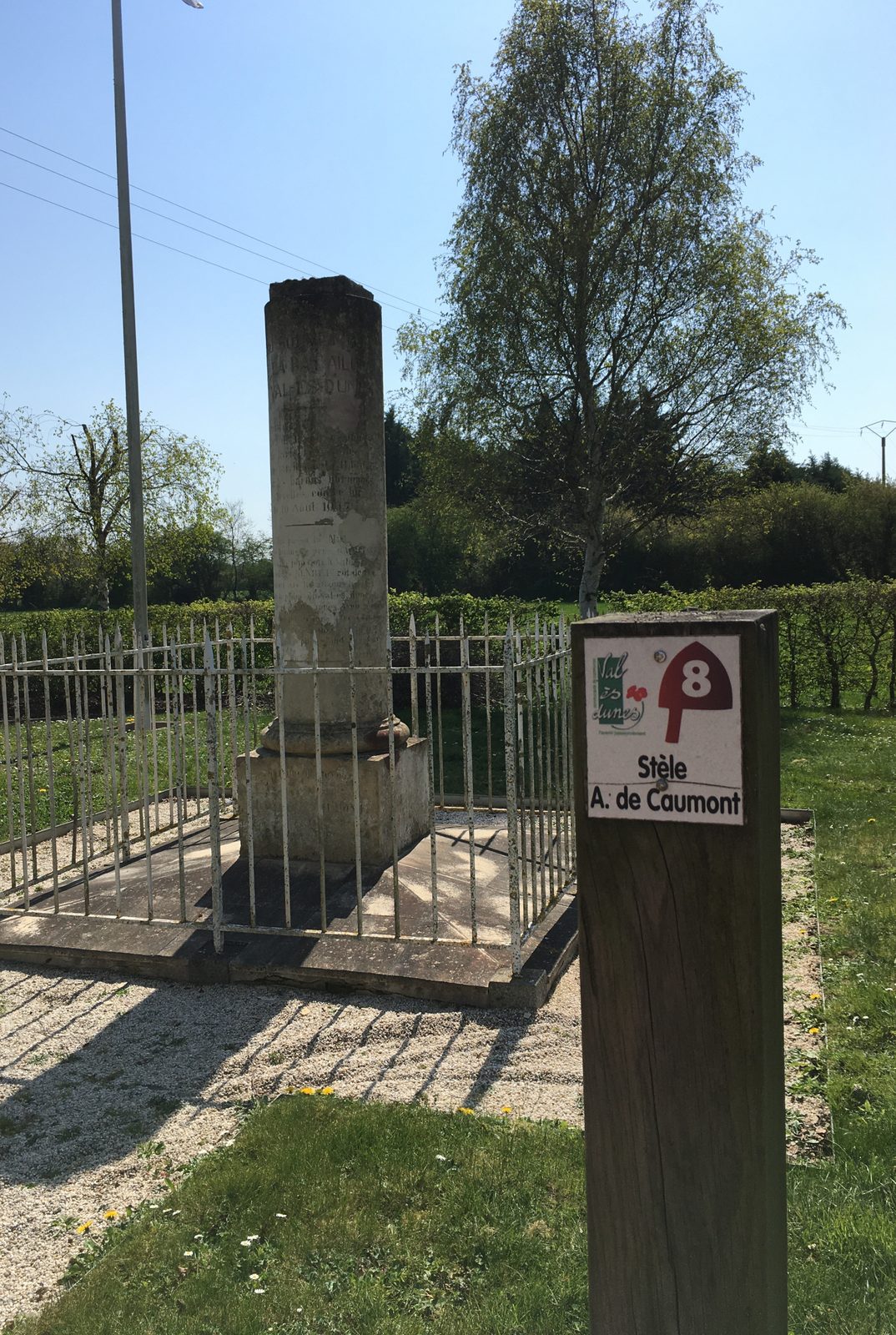 A stone column inside a small railed enclosure beside a marker post, under trees.