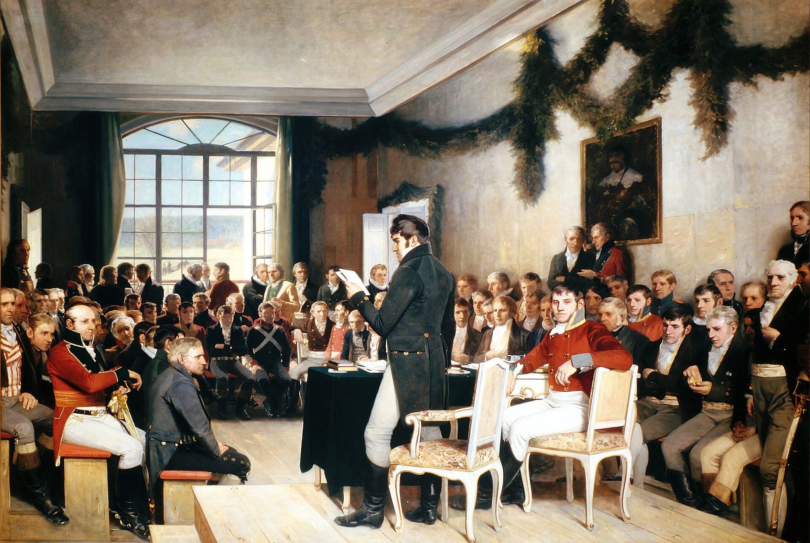 A painting of a crowded panelled room hung with green garlands, men in coats and uniforms seated on benches as one man stands to read.