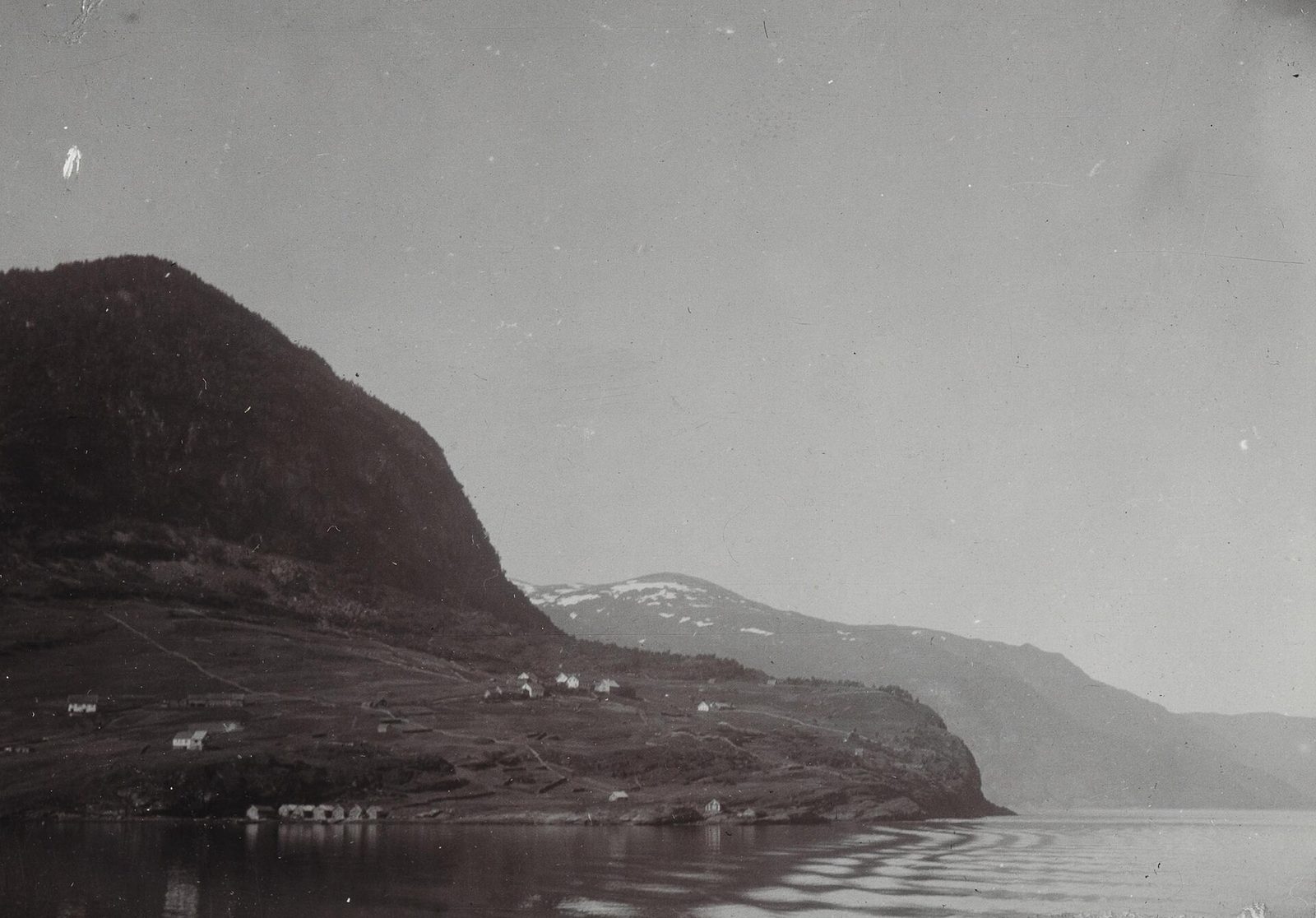 A black-and-white photograph of a steep wooded headland running down into still water, snow on the far mountains.