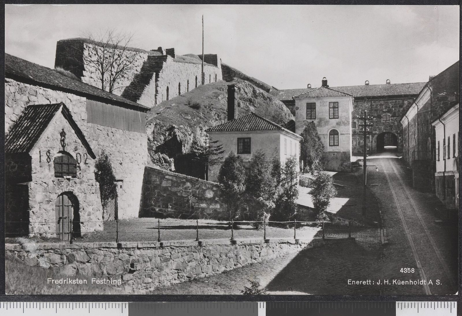 A black-and-white photograph of the inner yard of a fortress, stone walls and buildings around a cobbled lane.