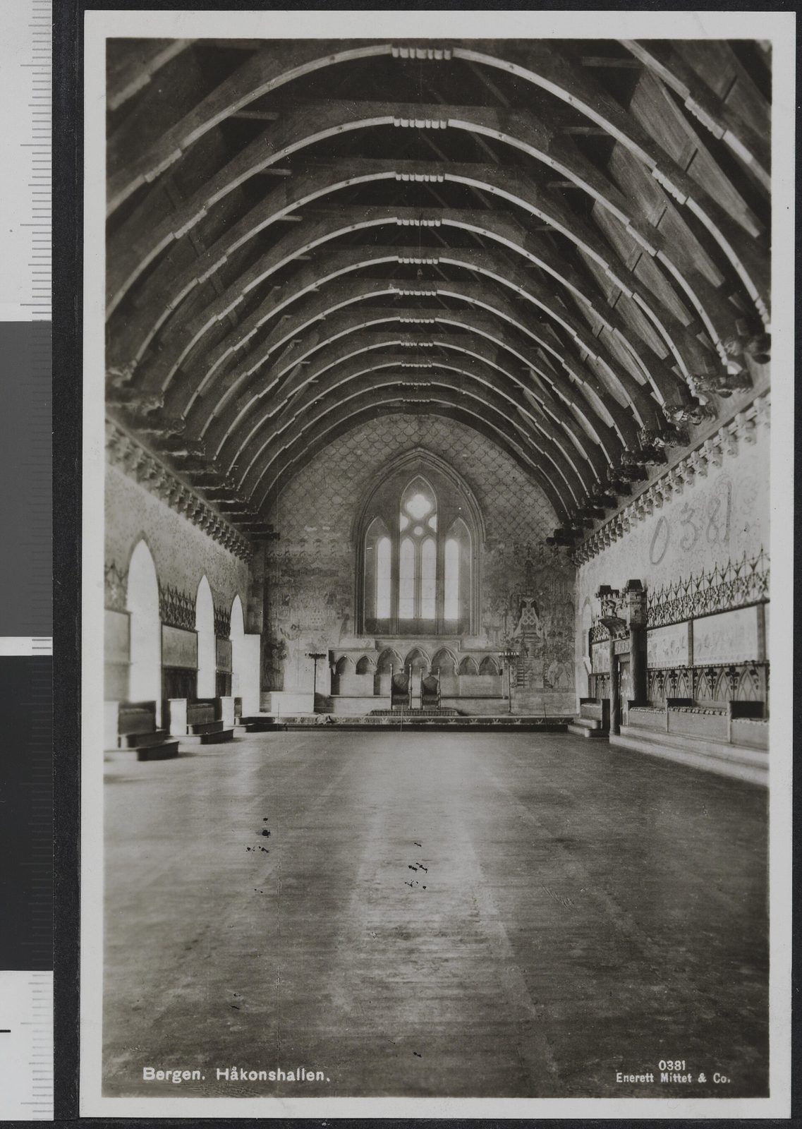 A black-and-white photograph of a long, empty stone hall with a timber roof and a tall Gothic window at the far end.