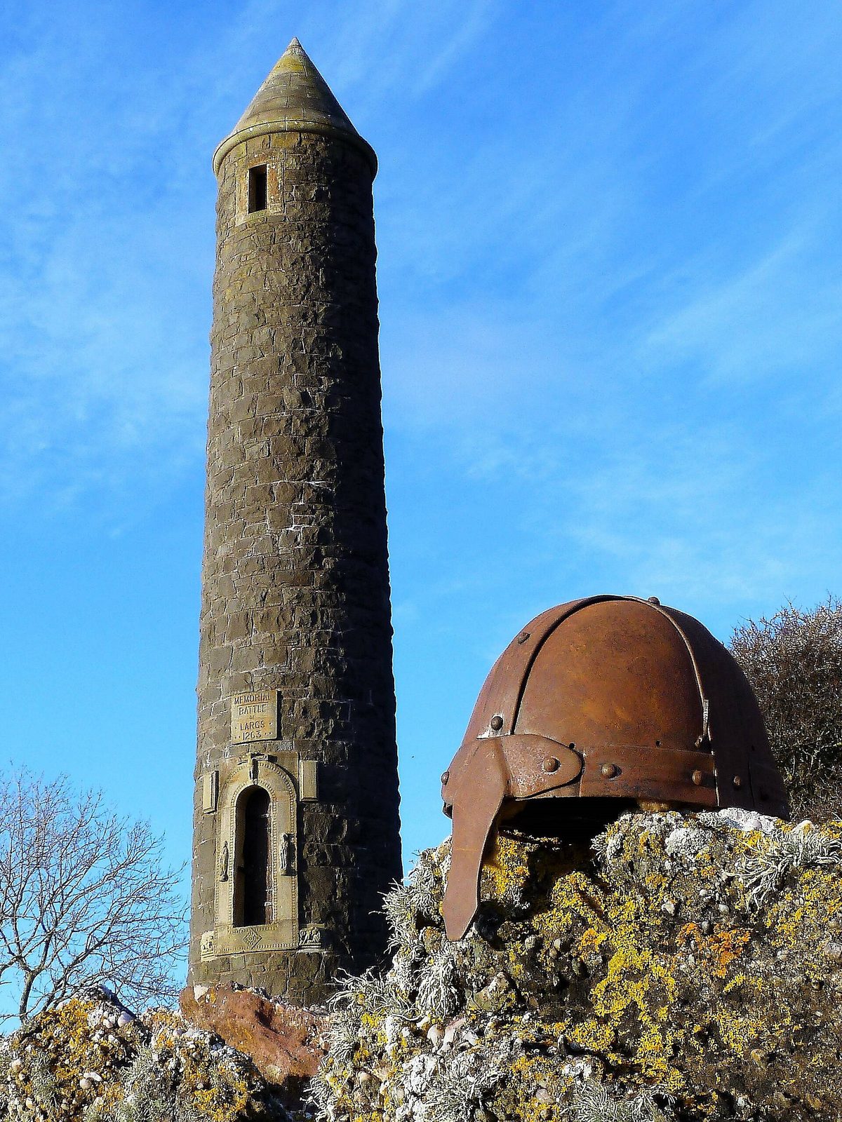 A tall round stone tower with a conical top standing on a rocky shore, a rusted iron helmet sculpture beside it.
