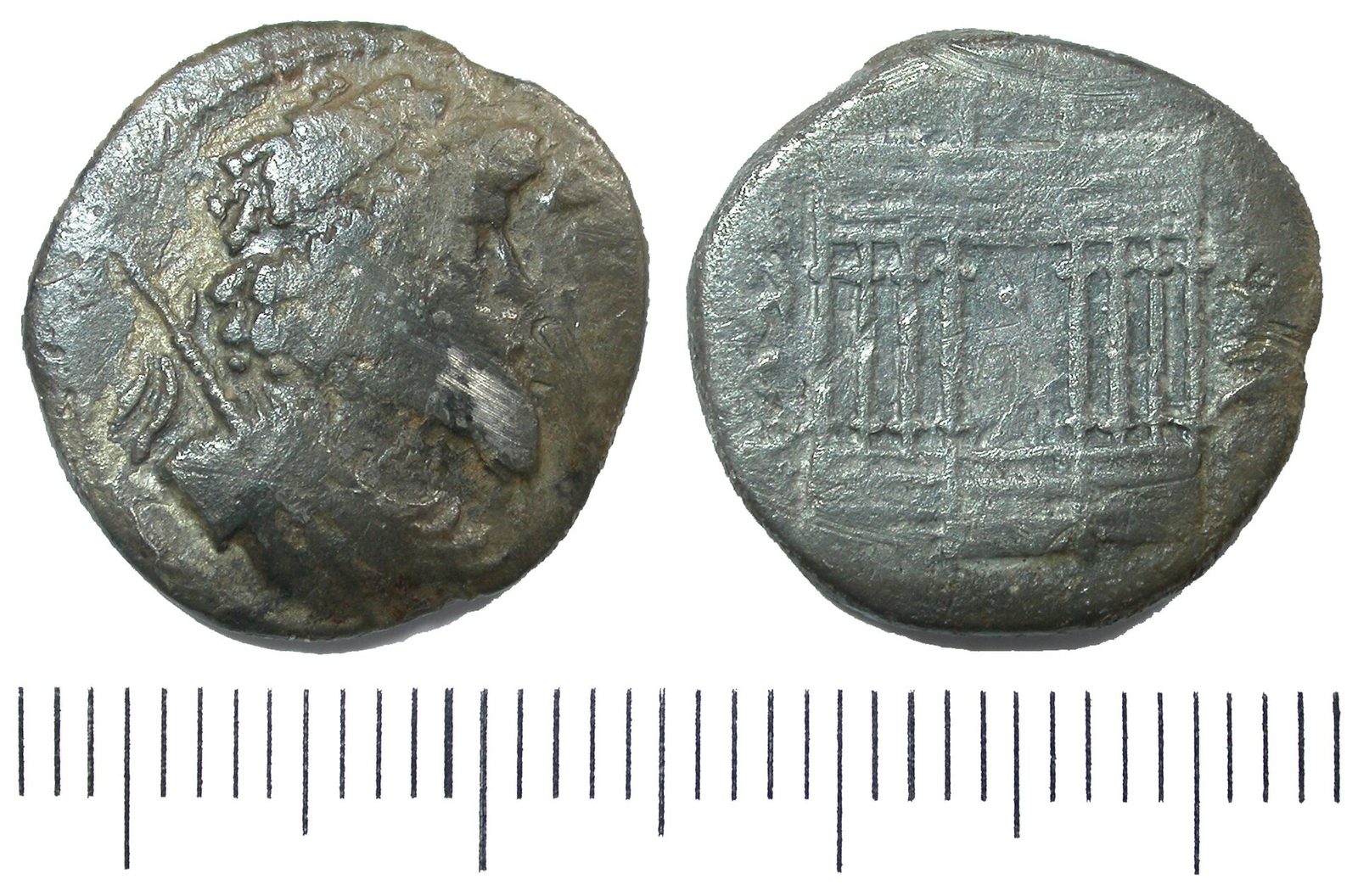 Both faces of a worn silver coin, a bearded bust and a columned temple, above a scale.