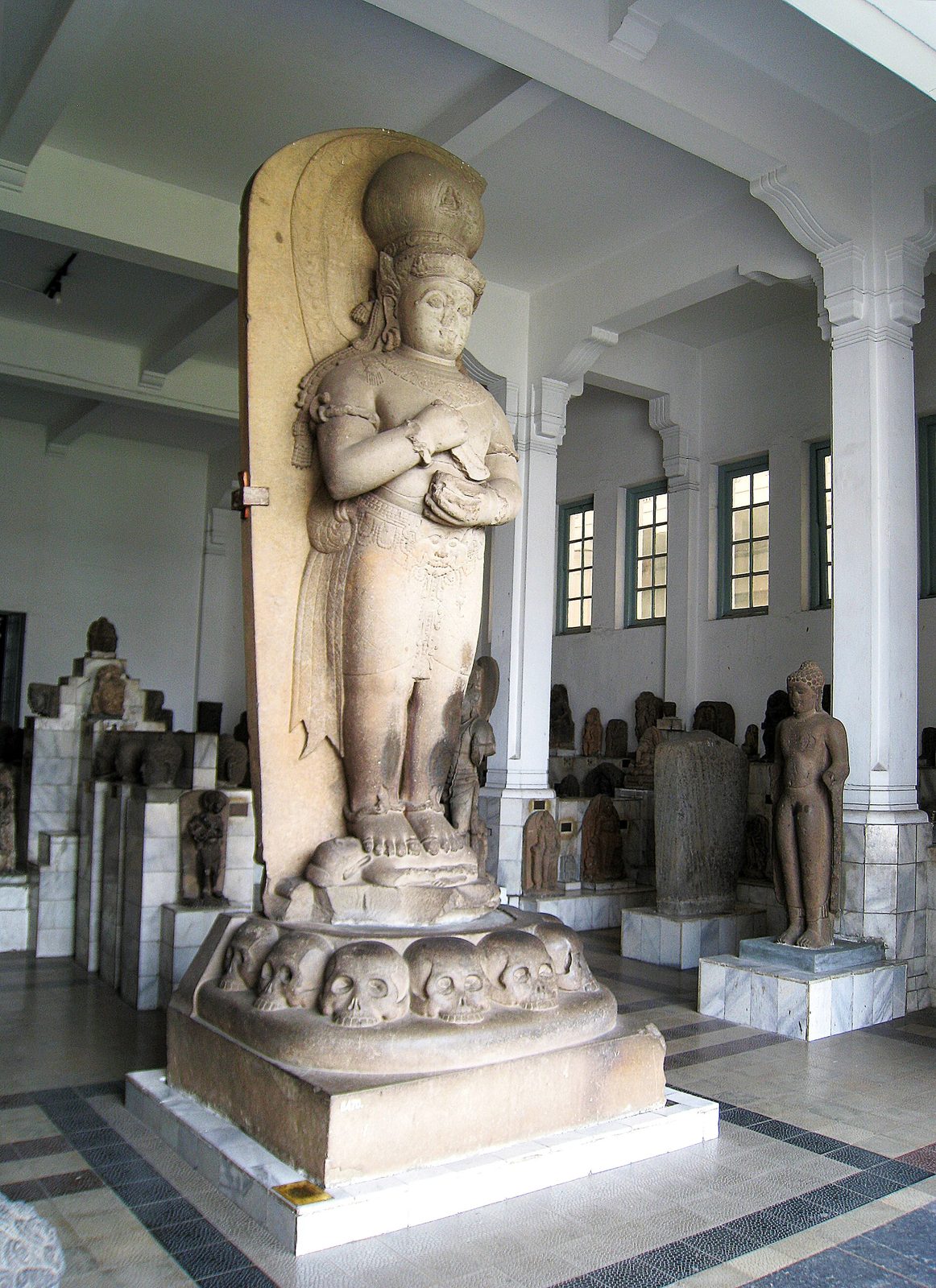 A colossal standing stone figure in a museum hall, towering over the smaller sculptures beside it, with a towering headdress, a knife held at the chest and a base carved with a row of skulls.